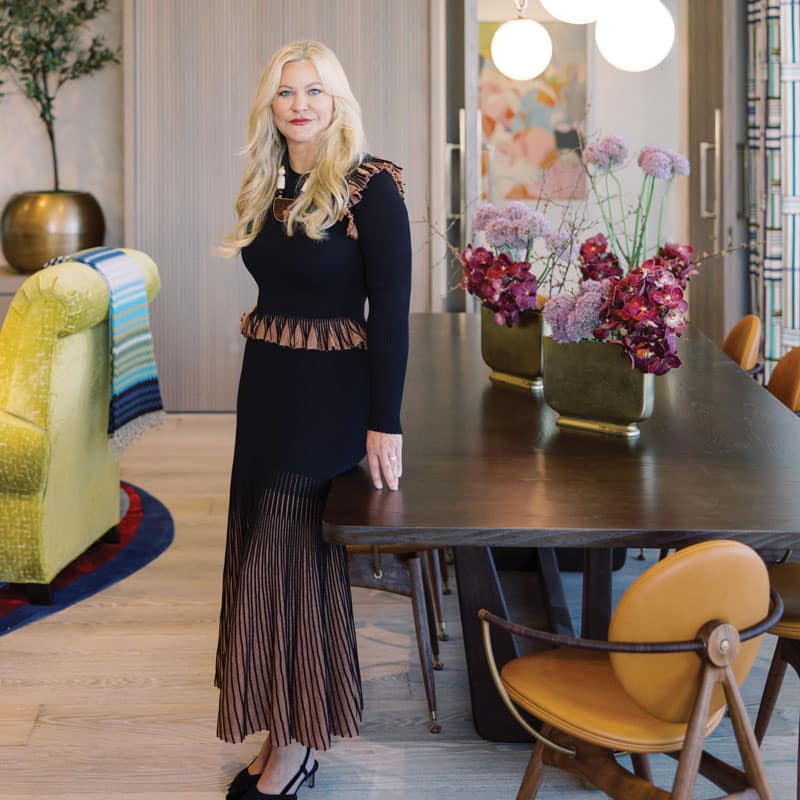 A woman in a black and brown dress stands by a wooden dining table with vases of pink flowers in a modern, well-lit living space.