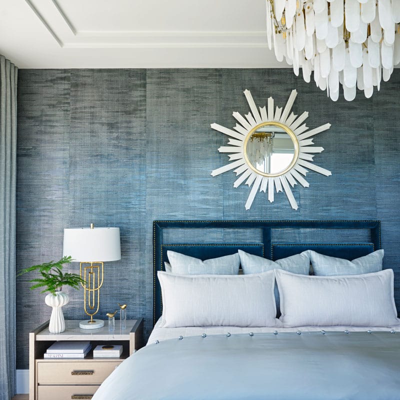 A neatly made bed with light blue bedding, a dark headboard, a sunburst mirror above, a nightstand with a lamp, books, and a vase of greenery, and a white chandelier overhead.