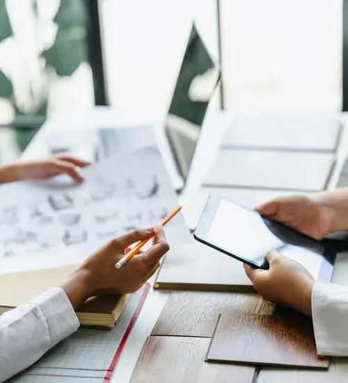 Two people sit at a table, one holding a tablet and the other holding architectural drawings and a pencil, with design samples spread out in front of them.