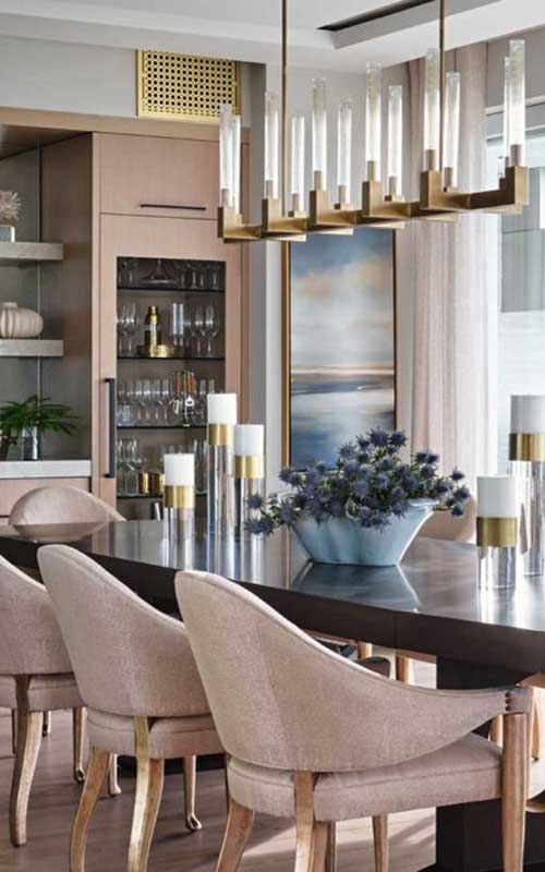 Modern dining room with a dark wood table, upholstered chairs, glass candle holders, a blue vase with flowers, and a contemporary chandelier. Shelves and art are visible in the background.