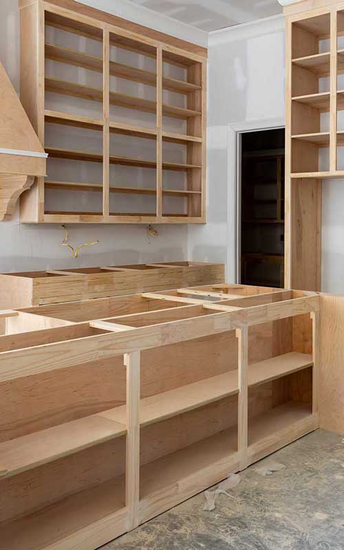 Unfinished wooden kitchen cabinets and shelves installed in a room under construction, with no doors, countertops, or appliances present.
