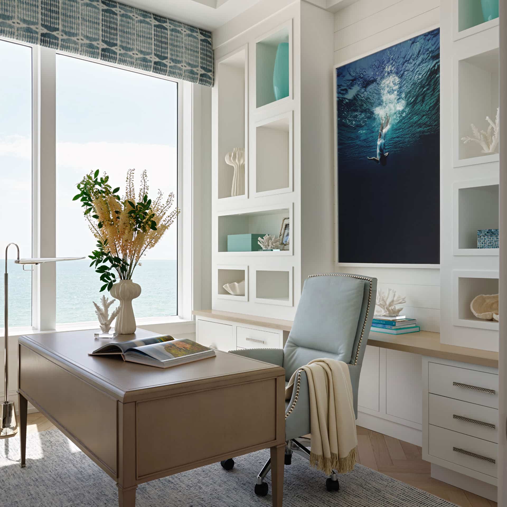 A sunlit home office with a desk, light blue chair, built-in shelves, ocean view window, and a large photo of a diver underwater on the wall.