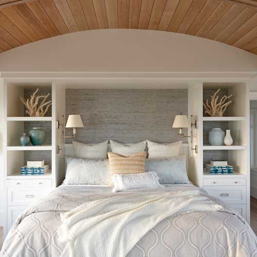 A neatly made bed with pillows and a beige blanket is set between built-in shelves holding vases, coral, and books. Wooden ceiling and soft lighting add warmth to the room.