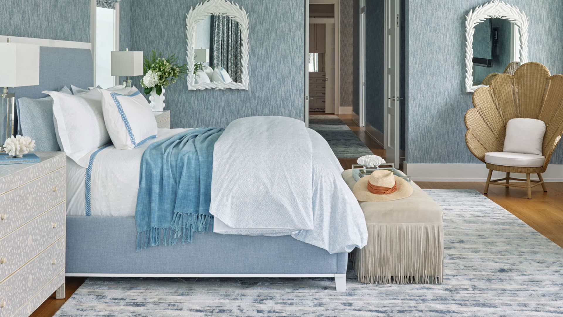 A neatly made bed with blue and white bedding in a stylish bedroom featuring mirrors, a fringed bench, and a peacock chair.
