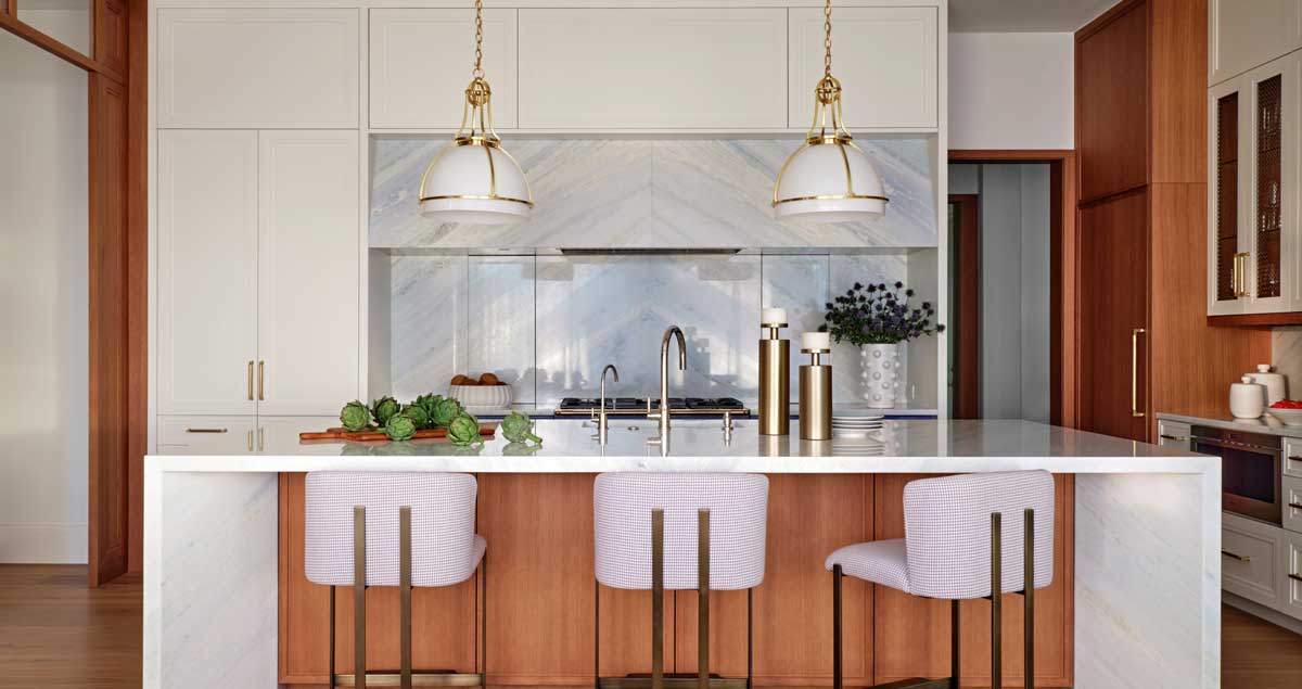 Modern kitchen with a marble island, three upholstered barstools, pendant lights, built-in appliances, and decorative plants on the counter.