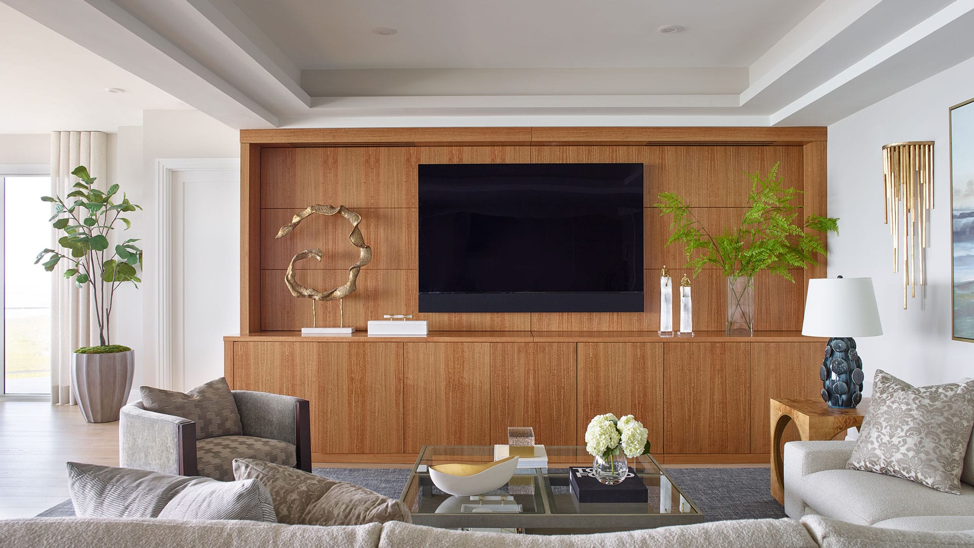 Modern living room with a large TV on a wooden wall unit, decorative objects, green plants, neutral-colored furniture, and a glass coffee table with flowers.