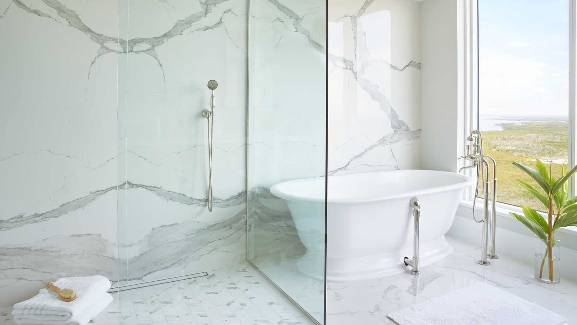 Modern bathroom with a white freestanding bathtub, glass-enclosed shower, marble walls and floor, a plant by the window, and folded towels on the floor.