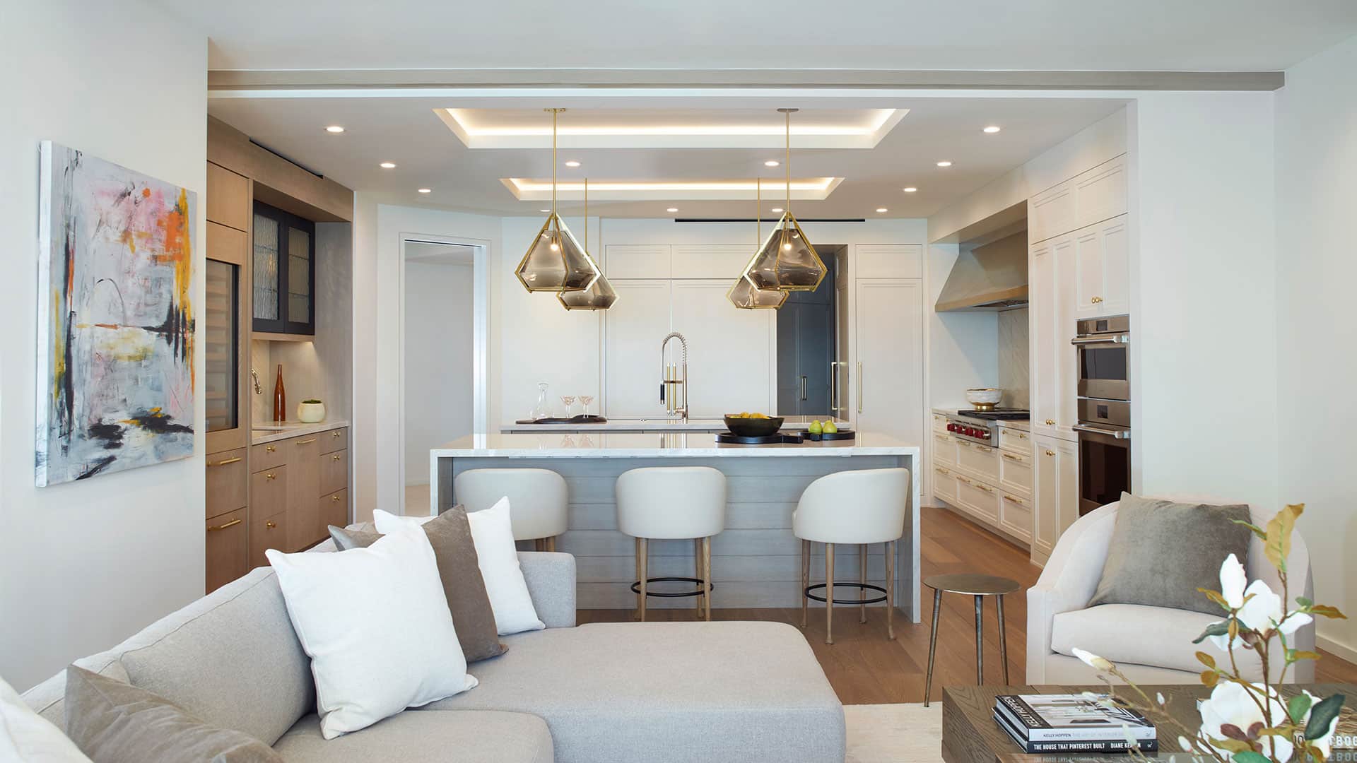 Modern kitchen with white cabinets, island seating, pendant lights, and stainless steel appliances. A neutral sofa and abstract painting are visible in the open living area.